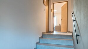 Eine Treppe führt zu einem Raum mit einem Bett und einem Sessel, einer Tür mit Griff und einem Lichtschalter an der Wand.