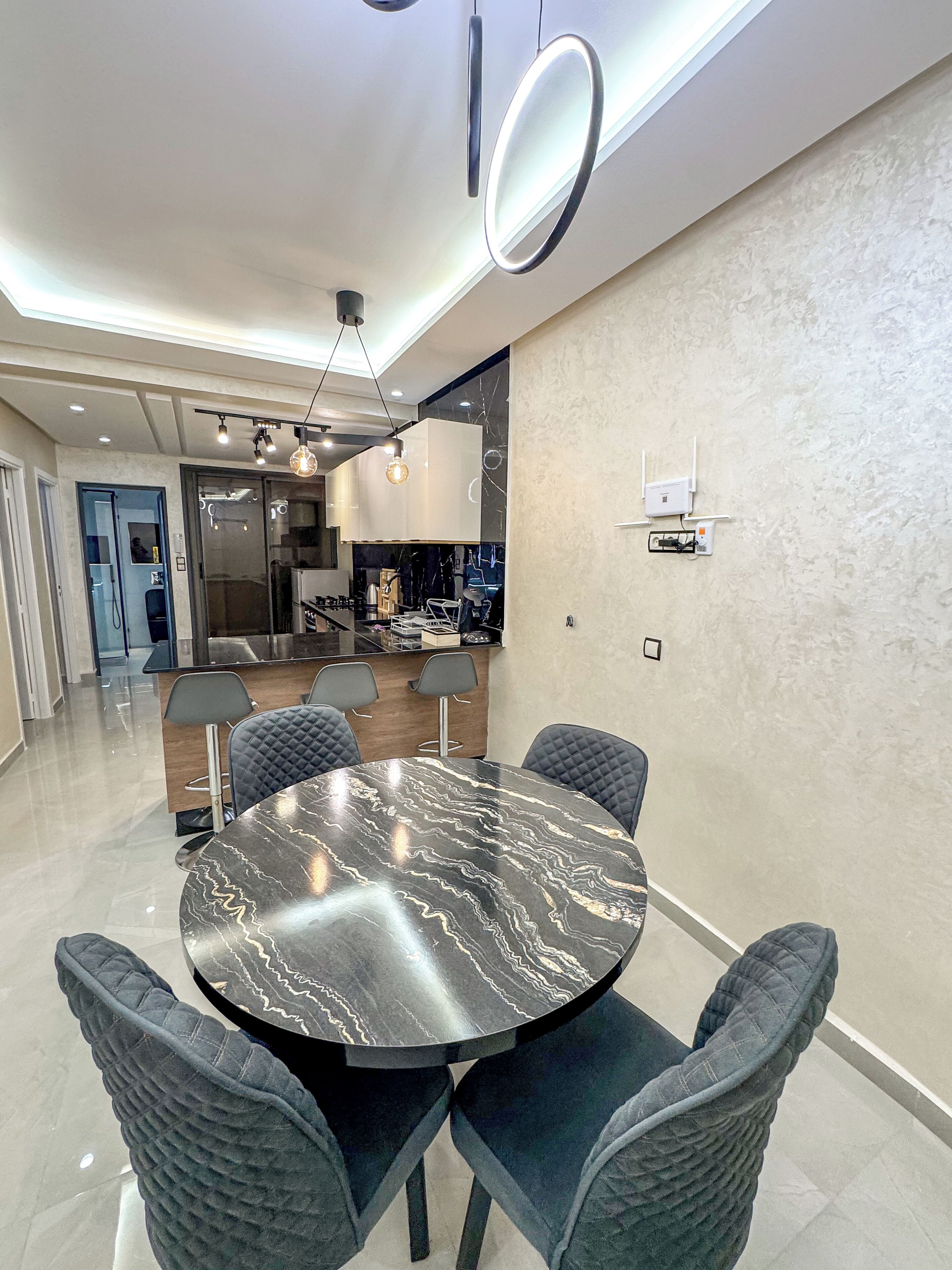 Un espace de salle à manger moderne avec une table en marbre, des chaises rembourrées, des luminaires suspendus et une cuisine en arrière-plan.