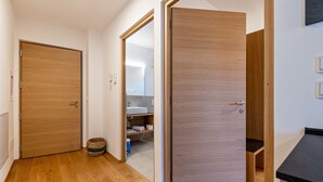 Um corredor com piso de madeira, uma porta fechada e uma porta aberta que dá para um banheiro com pia e prateleiras.