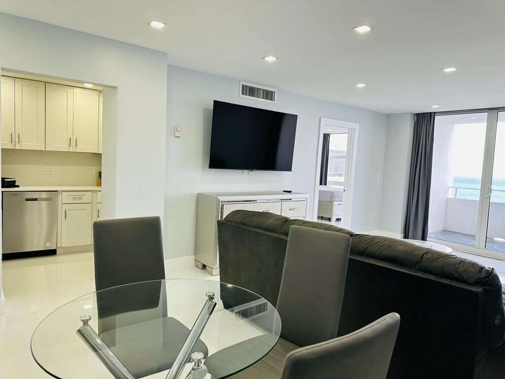Een moderne woonkamer met een glazen tafel, grijze stoelen, een bank, een flatscreen televisie en een keukenhoek met kasten en een vaatwasser.