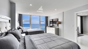 A modern bedroom with a large bed, a smaller bed, a TV, and a view of the sea.