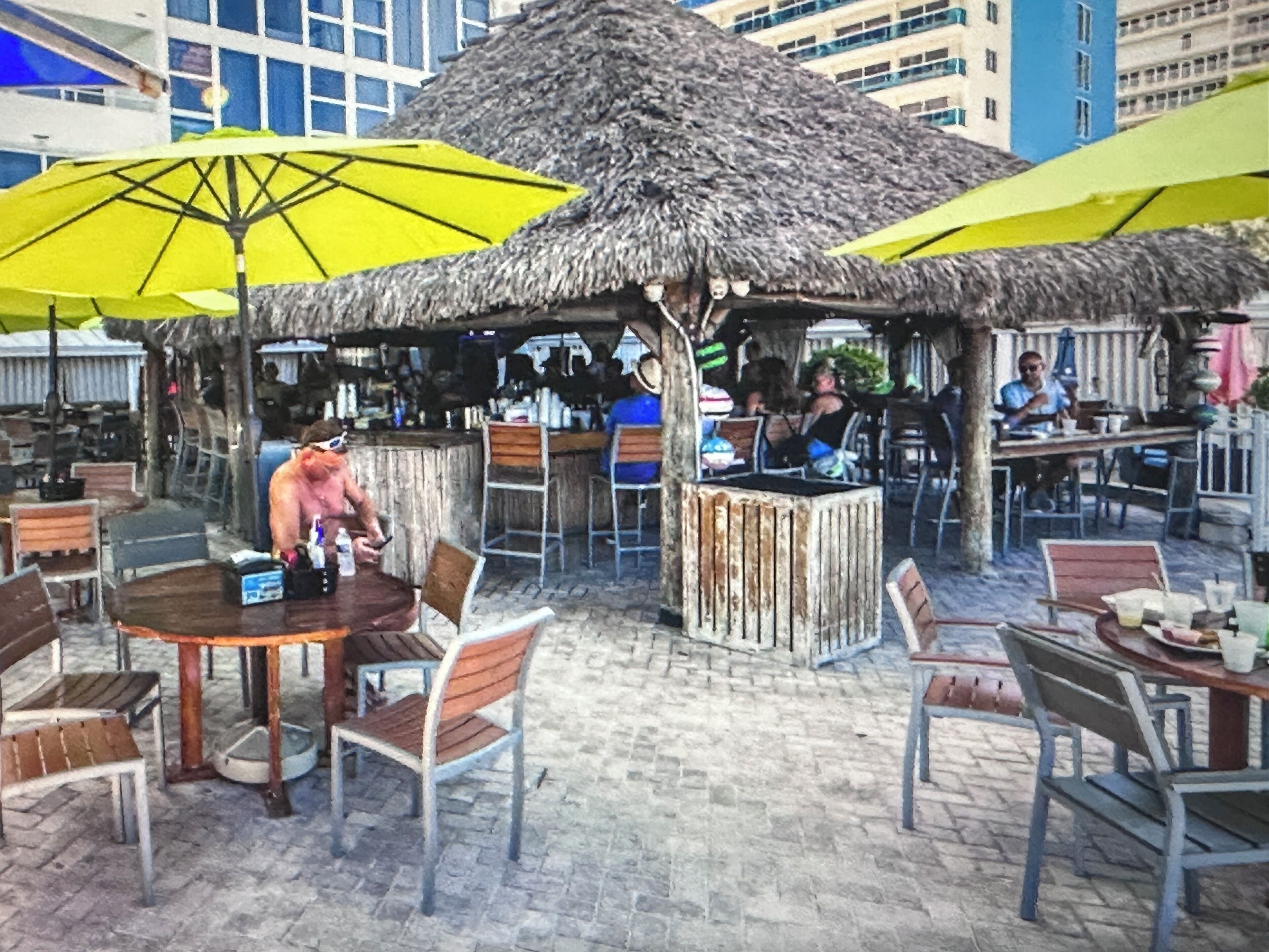 An outdoor dining area with thatched-roof tables and chairs, surrounded by buildings.