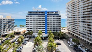 A hotel with a blue and white facade, multiple floors, and a parking lot with cars and palm trees.
