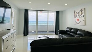 A modern living room with a black leather sofa, a flat-screen TV, and a large window offering a view of the sea.