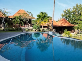 A pool area with a clear blue pool, surrounded by tropical plants and buildings with red-tiled roofs.