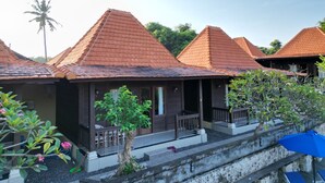 A row of bungalow-style accommodations with sloped roofs, wooden balconies, and a garden area with potted plants and a palm tree. - Villa Vilah (Penida Island)
