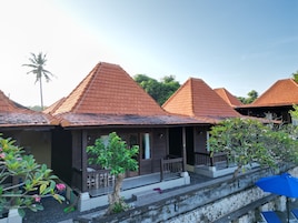 A row of bungalow-style accommodations with sloped roofs, wooden balconies, and a garden area with potted plants and a palm tree.