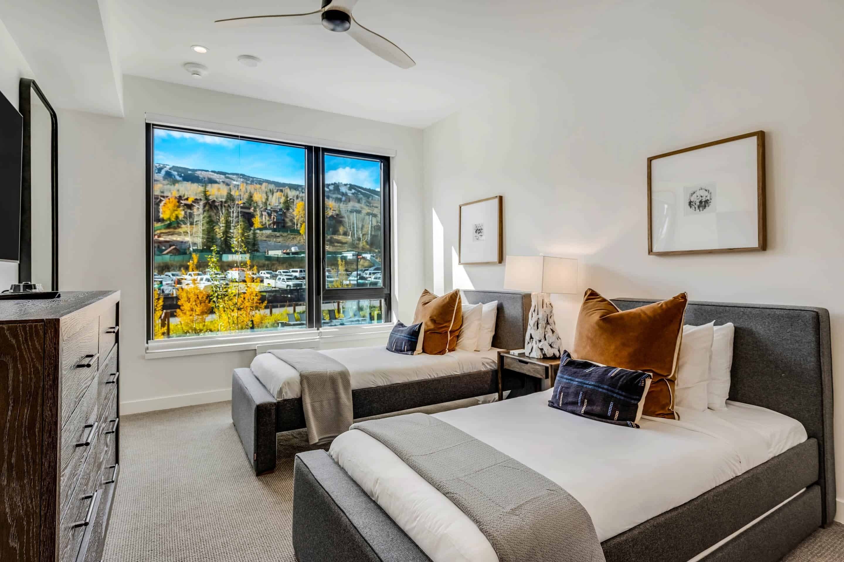 Ett hotellrum med två sängar, ett stort fönster med utsikt över bergen, en takfläkt och inramade tavlor på väggen.