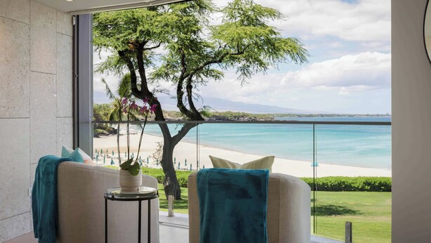 Une chambre moderne dotée d’une grande fenêtre donnant sur une plage et l’océan. On y trouve deux canapés agrémentés de coussins turquoise, ainsi qu’une petite table d’appoint sur laquelle est posée une plante.