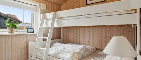 A bunk bed room with a ladder, a bedside lamp, and a window with a view of a house and a plant.