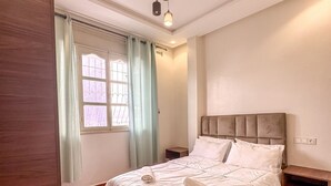 A hotel room with a bed, a nightstand, a window with curtains, and a ceiling light fixture. - El-wifaq - Appartement 2 (Agadir)