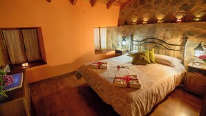 A bedroom with a stone wall, wooden ceiling beams, a bed with towels, a nightstand, and a window with curtains. - Casa Rural en la Montaña de León. Entre Valles (Benllera)