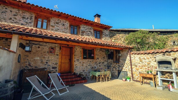 A stone building with a tiled roof, a wooden door, and a brick chimney. There are two outdoor tables with chairs, a small garden, and a stone wall. - Casa Rural en la Montaña de León. Entre Valles (Benllera)