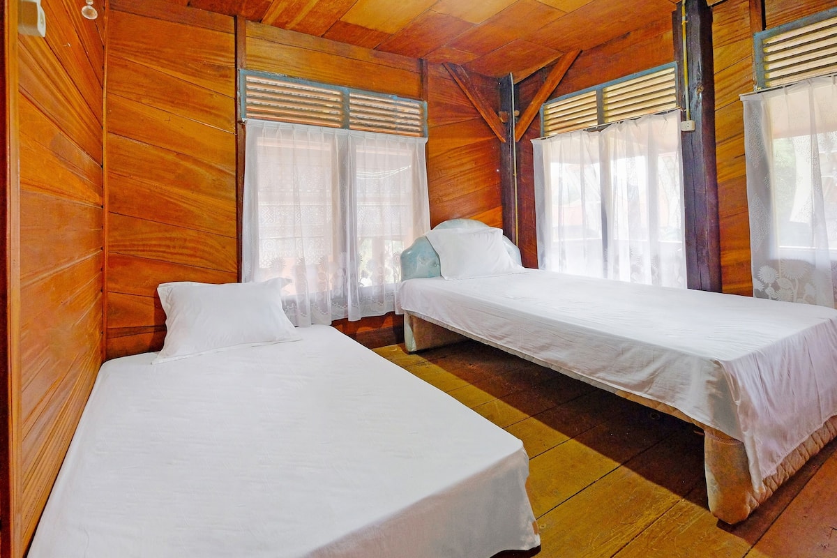 Una habitación con dos camas individuales, paredes de madera y ventanas grandes con persianas.