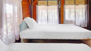 Uma cama de solteiro com roupa de cama branca num quarto com paredes de madeira e uma janela com cortinas leves.