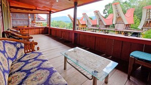 Uma área de estar coberta ao ar livre, com mobiliário de madeira, uma mesa de vidro e vista para edifícios tradicionais.