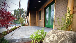 Un bâtiment en bois avec une porte et des fenêtres vertes, un chemin pavé et un petit jardin agrémenté de plantes et d’un rocher.