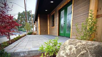 A wooden building with a green door and windows, a paved walkway, and a small garden area with plants and a rock.