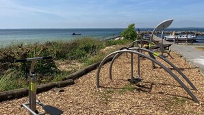 Ein Fitnessbereich direkt am Strand mit Metallgeräten, einer Holzterrasse und Blick auf das Meer und Boote.