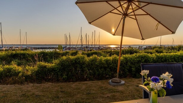 Ein Tisch mit Sonnenschirm, einer Blumenvase und Sonnenbrille, mit Blick auf einen Hafen voller Boote und einem Sonnenuntergang im Hintergrund.