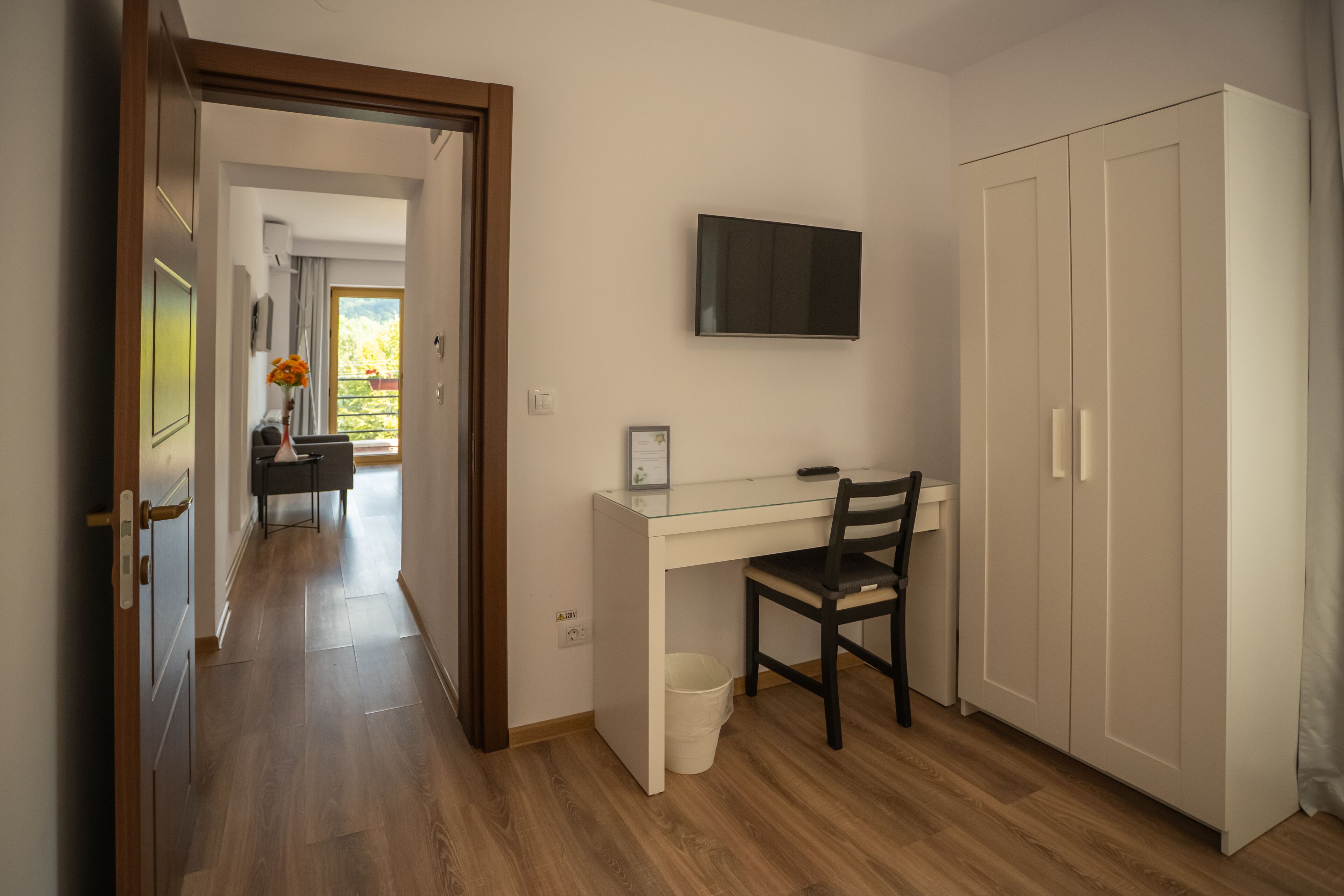 Quarto com escrivaninha, cadeira, televisão, guarda-roupa e corredor que dá para outro quarto.