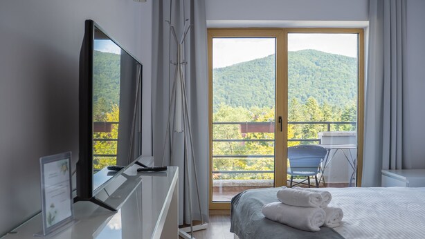 Una habitación de hotel moderna con una cama, un escritorio con un televisor de pantalla plana y un balcón con vistas a colinas verdes.