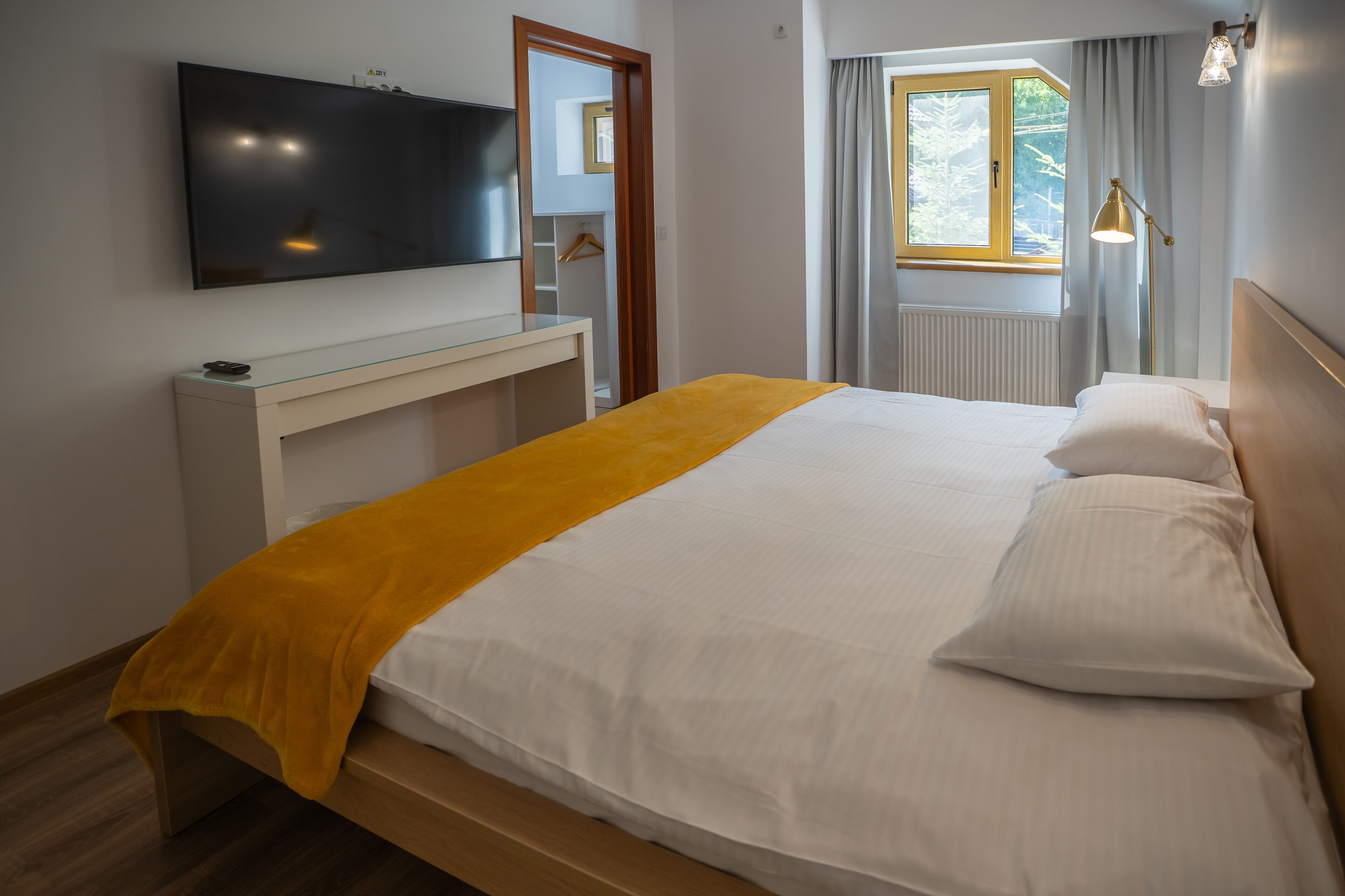 A modern hotel room with a large bed, a TV mounted on the wall, a bedside table, and a window with curtains.