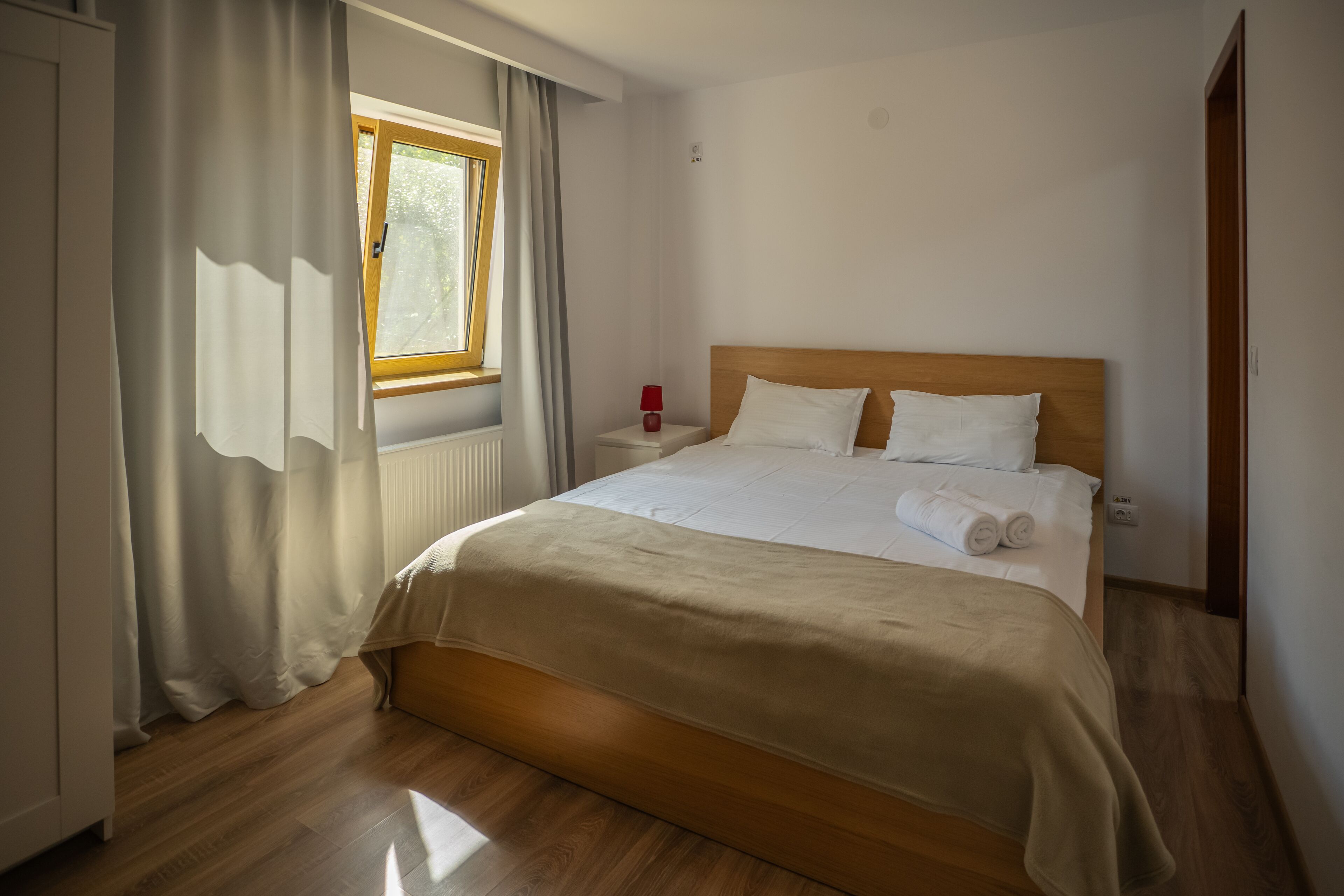 A neatly made bed with a wooden headboard, a window with curtains, and a bedside table with a lamp.
