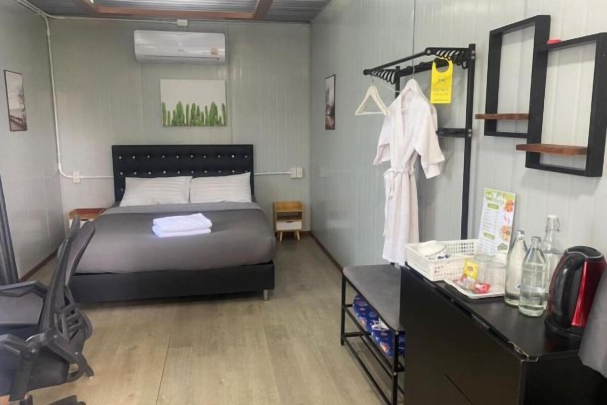 A hotel room with a bed, a desk with a kettle, a chair, a small table, and a wall-mounted drying rack.