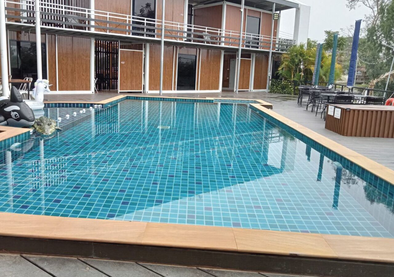 Piscina retangular com azulejos azuis, rodeada por um deck de madeira e mobiliário para áreas externas.