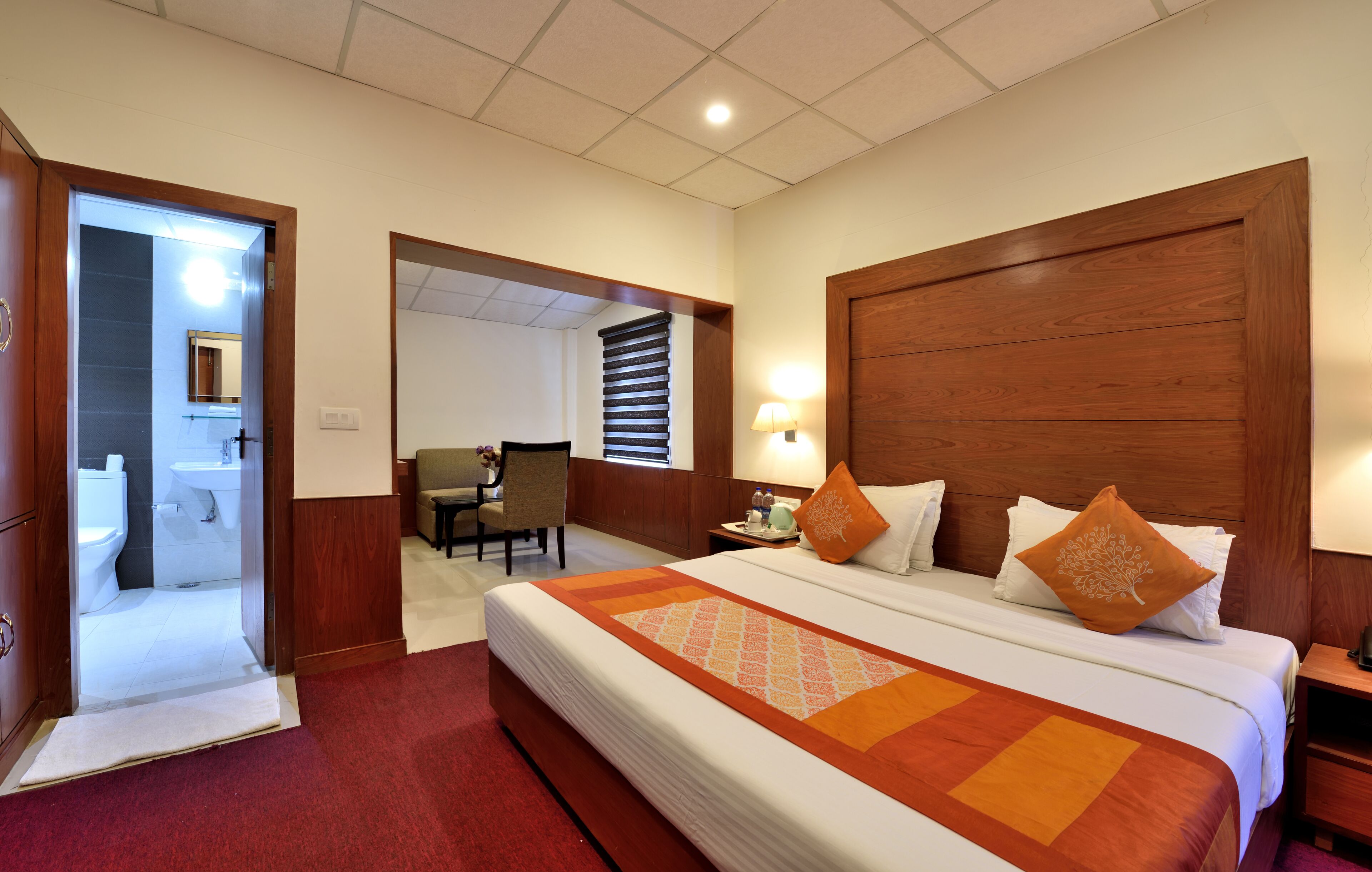 A hotel room with a large bed, a wooden headboard, a nightstand, a chair, a desk, and a bathroom visible through an open door.
