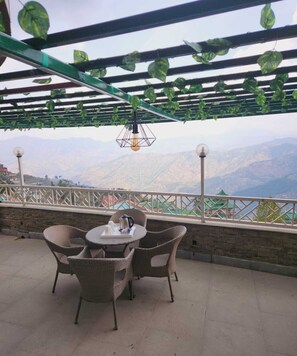 A terrace with wicker furniture and a table set for two, overlooking a mountainous landscape. - Galleu Hill Resort, Kufri, Shimla By LA Riqueza Hotels (Theog)