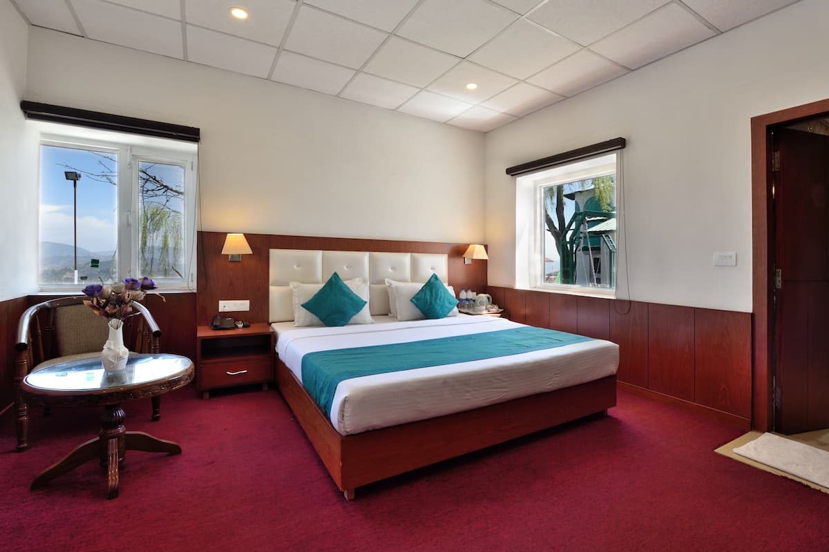 Habitación de hotel con una cama grande, dos mesitas de noche, una mesita redonda con un florero y dos ventanas que dan al exterior.