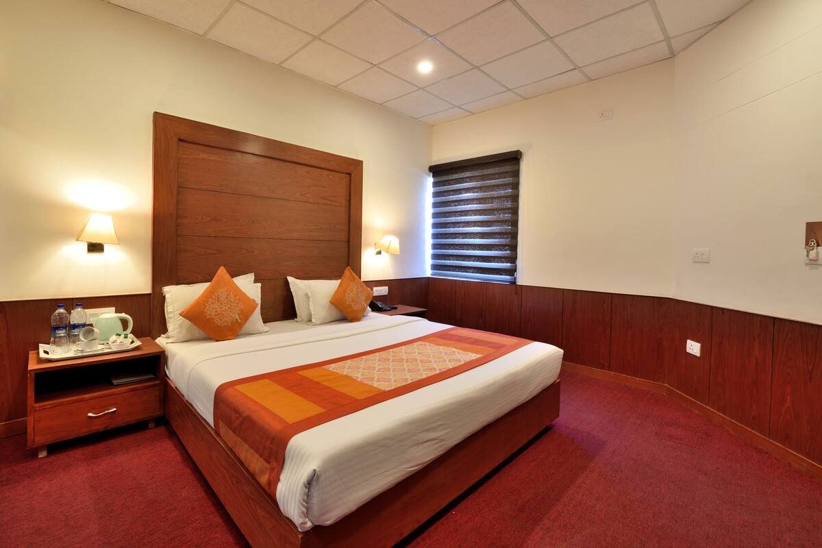 Habitación de hotel con cabecera de madera, cama con ropa blanca y almohadas naranjas, mesita de noche con lámpara, alfombra roja y ventana con persianas.