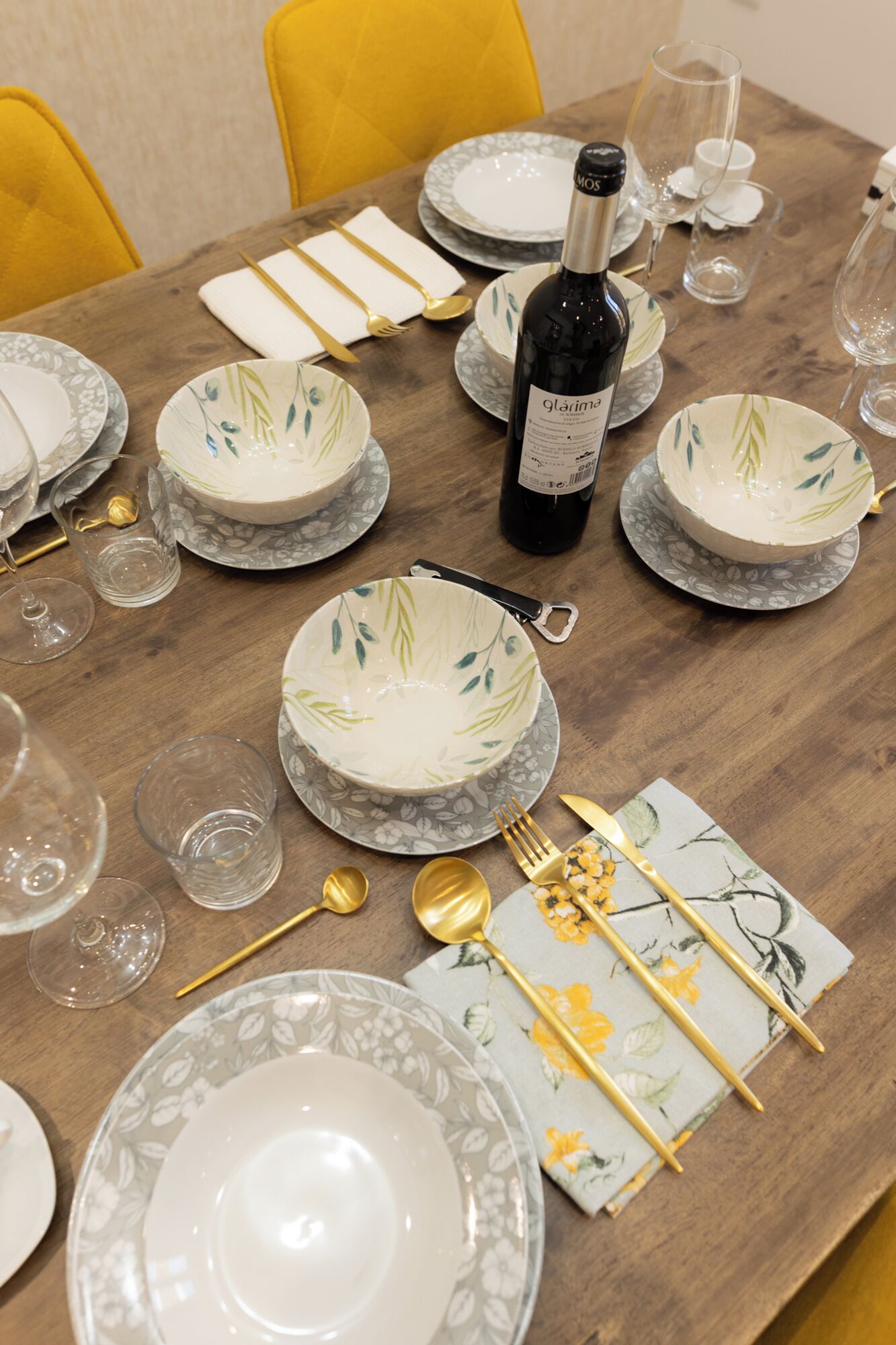 A dining table set with floral-patterned plates, bowls, and cutlery. A bottle of wine is placed on the table.