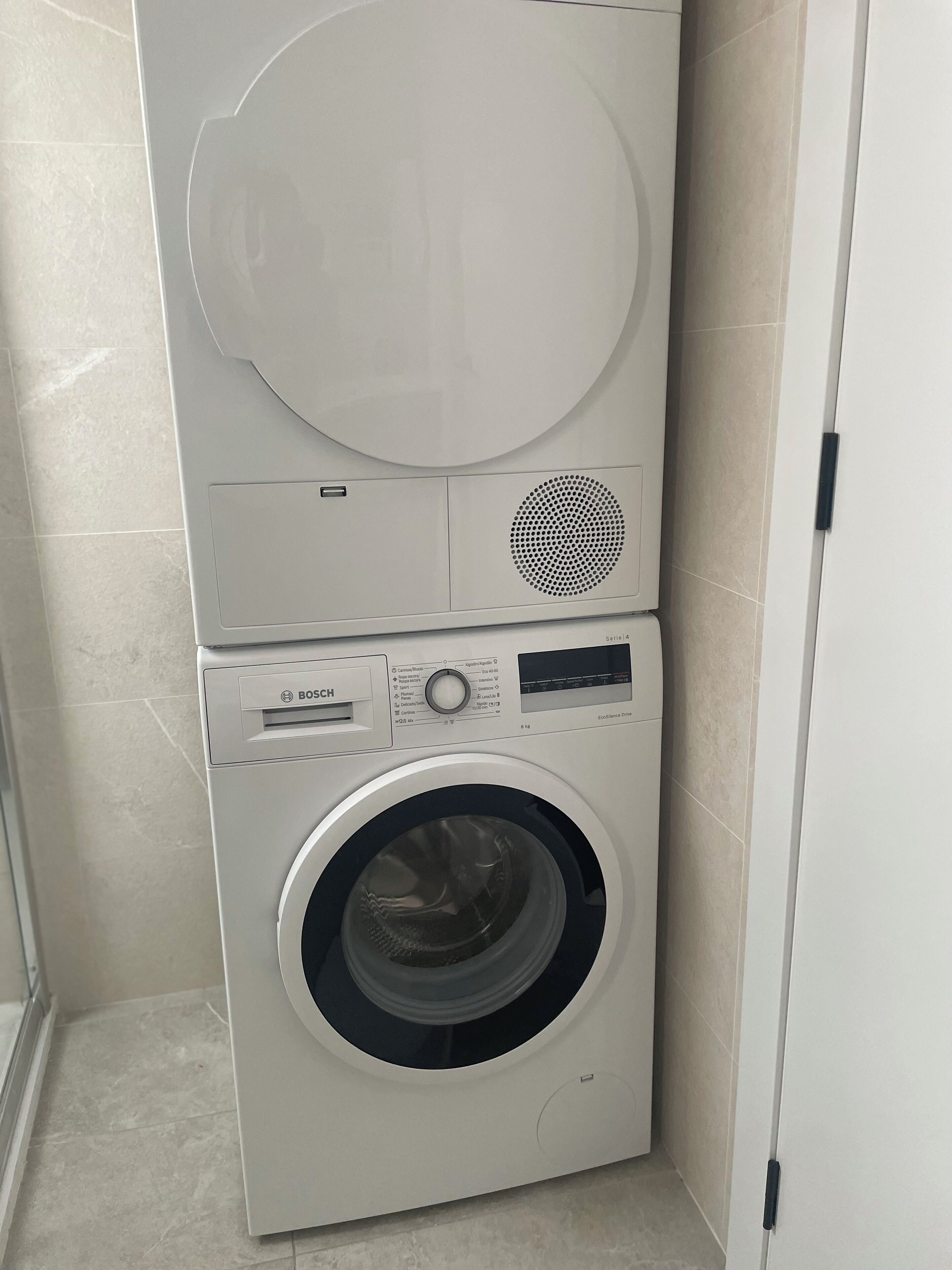 A compact, white Bosch washing machine and dryer unit in a corner of a room with tiled walls and floor.