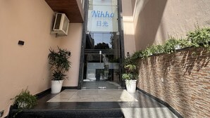 Ein Gebäudeeingang mit Glastür, einem Schild mit der Aufschrift 'Nikko' und je einer Pflanzengruppe auf jeder Seite.