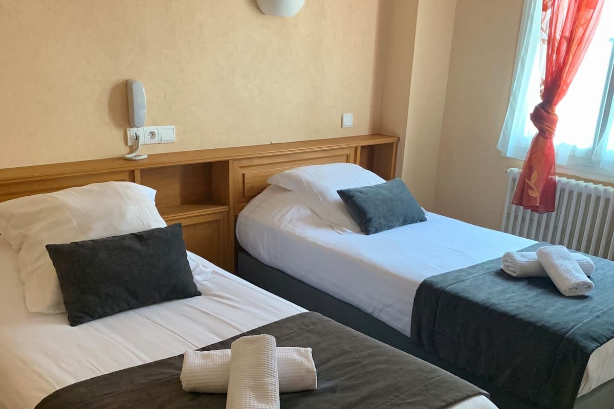 Quarto de hotel com duas camas, cada uma com roupa de cama branca e cabeceiras escuras. Há duas mesas de cabeceira com abajures, uma janela com cortinas vermelhas e um radiador.