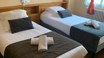 Habitación de hotel con dos camas, cada una con sábanas blancas y cabeceros oscuros. Hay dos mesitas de noche con lámparas, una ventana con cortinas rojas y un radiador.