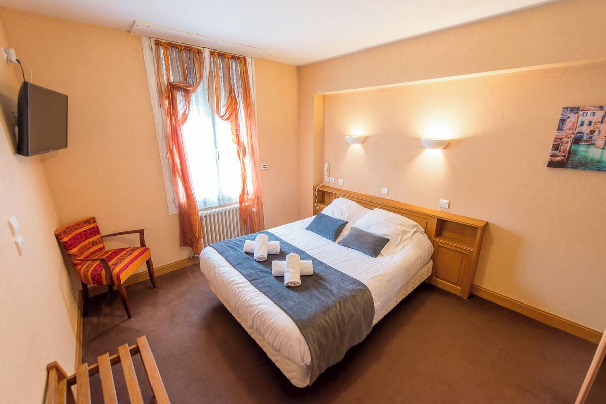 Quarto de hotel com cama, uma cadeira, radiador e um quadro na parede.