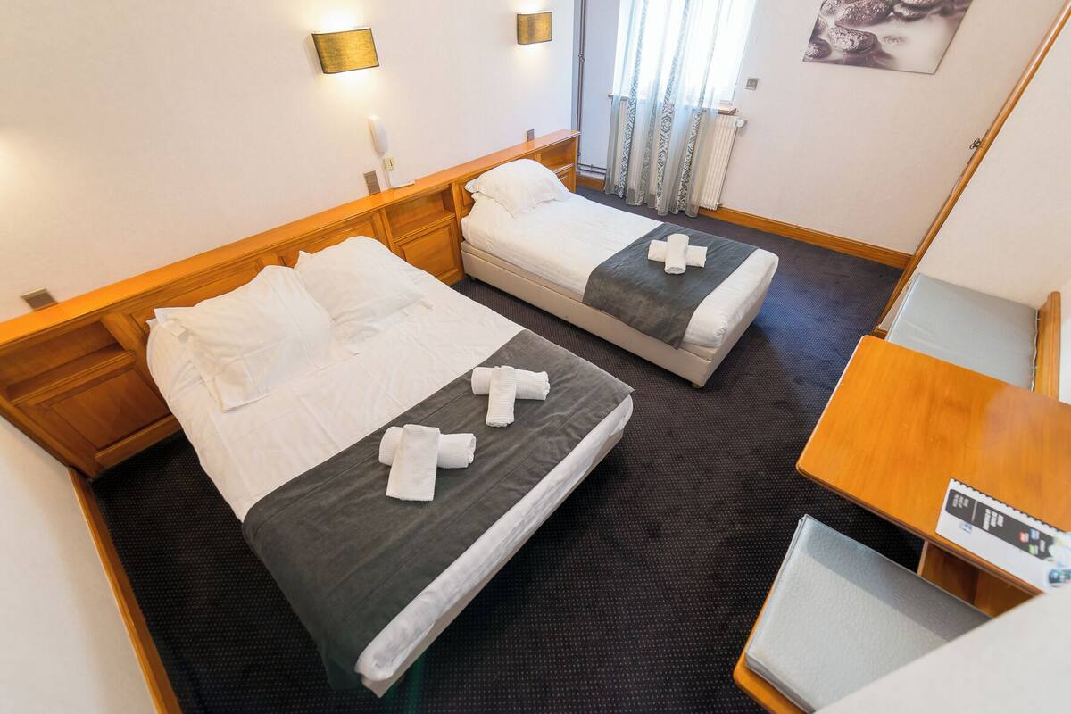 Quarto de hotel com duas camas, cabeceira de madeira, uma escrivaninha e uma janela com cortinas.