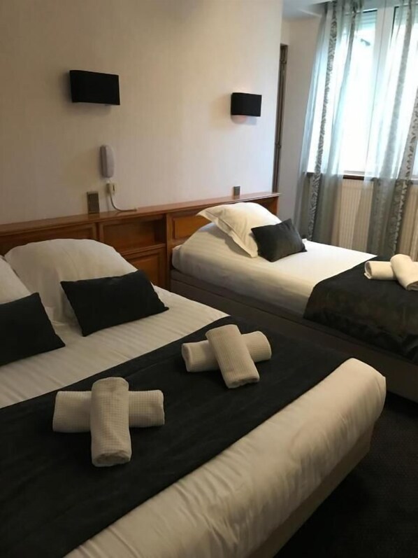 Habitación de hotel con dos camas, cada una con ropa blanca y almohadas oscuras. Hay dos mesitas de noche con lámparas, un televisor de pantalla plana montado en la pared y una ventana con cortinas.
