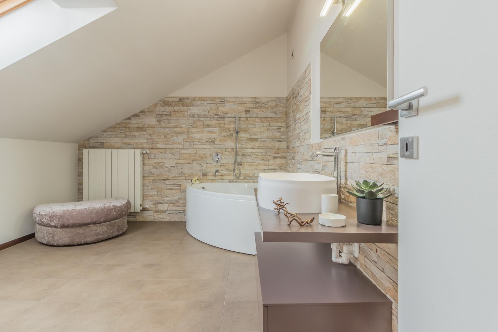 A modern bathroom with a stone wall, a freestanding bathtub, a vanity with a mirror, and a small potted plant.