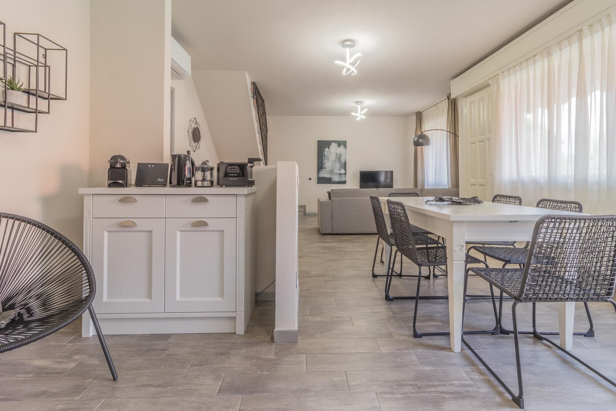 Una cocina moderna con armarios blancos, un comedor con mesa blanca y sillas negras, y un amplio ventanal con cortinas traslúcidas.