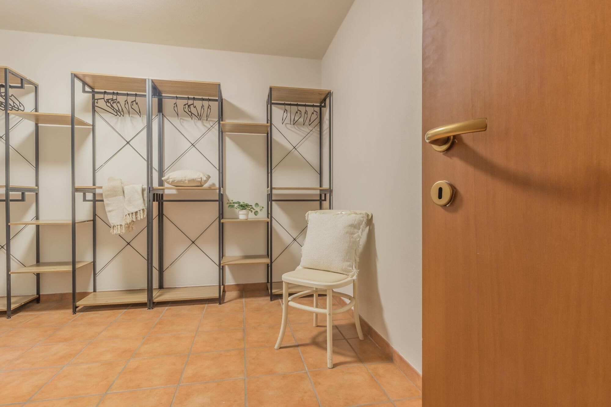 Um quarto com porta de madeira, uma cadeira branca e três prateleiras de metal com diversos objetos.