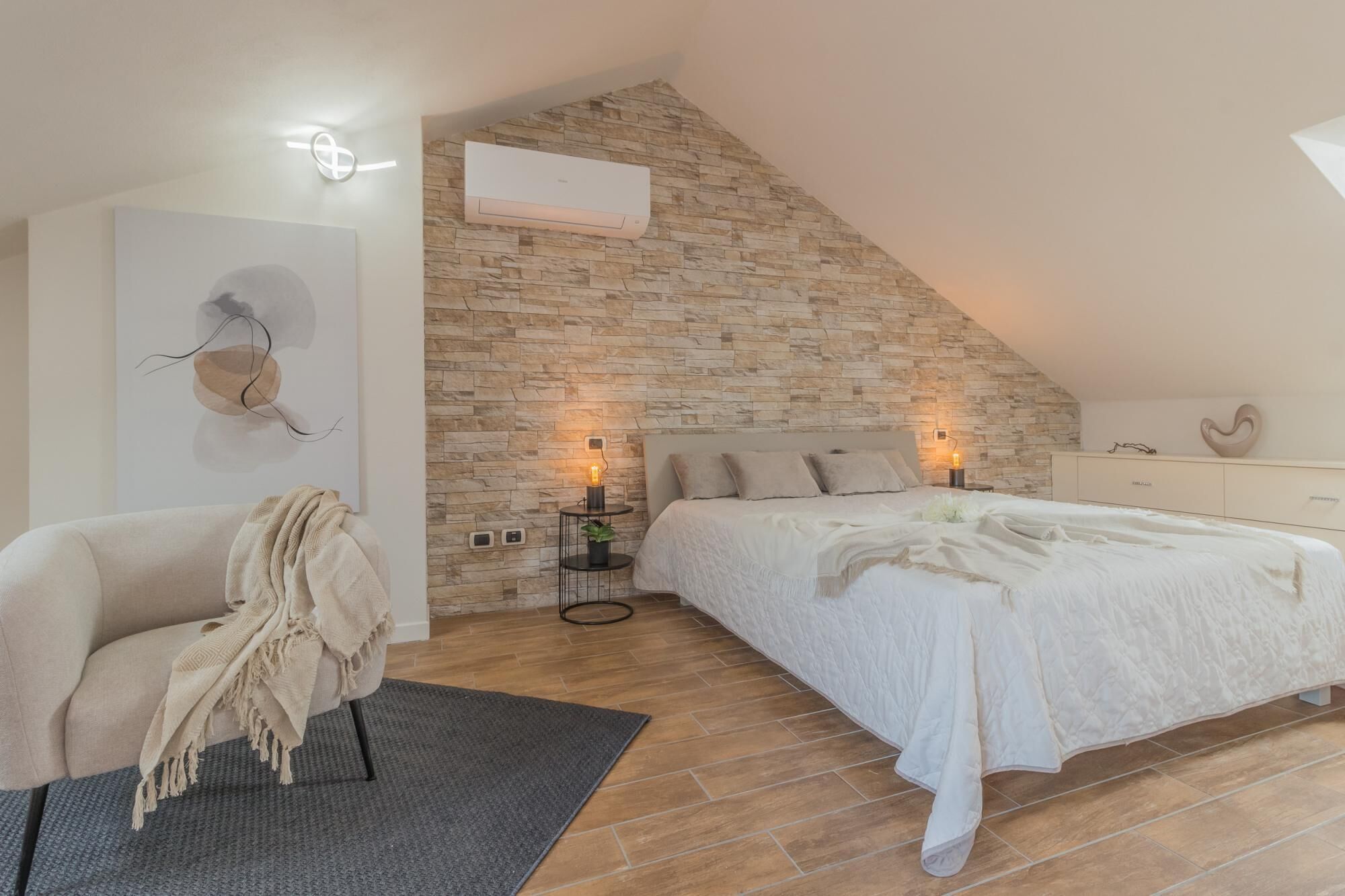 A bedroom with a sloped ceiling, stone wall, a bed with white bedding, a bedside table with a lamp, a chair with a blanket, and a wall-mounted air conditioner.