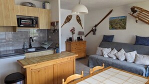Una cocina compacta con microondas, pileta y armarios de madera. Cuenta con una mesa de comedor con sillas, un basurero pequeño y una cama con almohadas y una colcha.