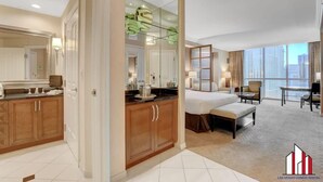 Quarto de hotel moderno com uma cama grande, área de estar com uma poltrona e mesa, cozinha compacta e uma janela ampla com vista para a cidade.