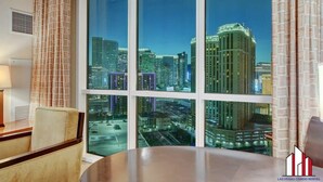 Quarto de hotel com uma grande janela com vista para a paisagem urbana, uma mesa redonda e uma cadeira.