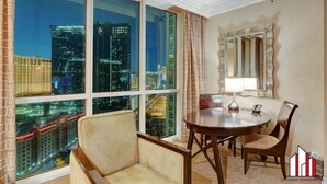 Quarto de hotel com uma janela grande que oferece vista da cidade, uma mesa de jantar redonda com cadeiras, um espelho e um abajur.